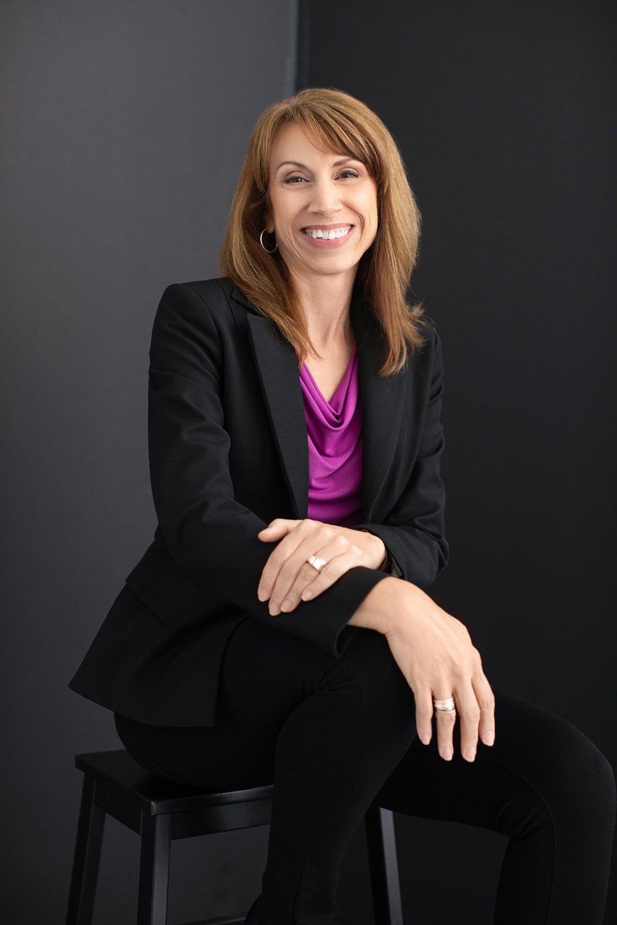 Woman in a black suit and purple shirt sitting on a stool against a dark background
