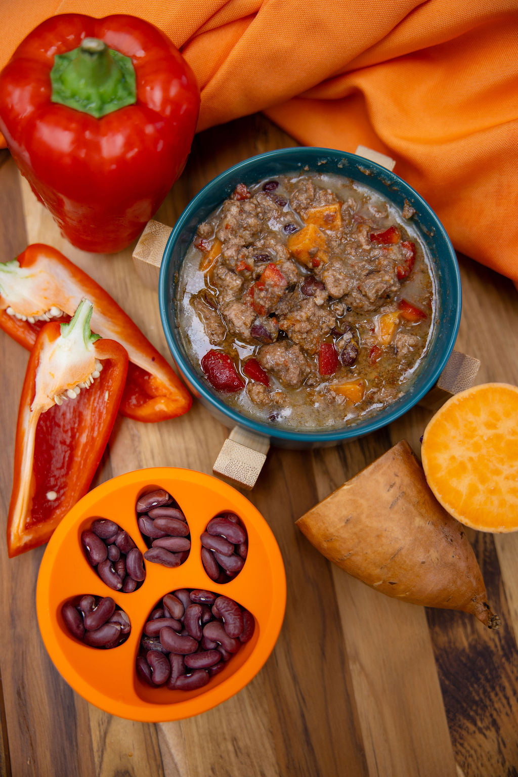 Assorted food items including a red pepper, beans, a bowl of soup, and sweet potatoes on a wooden surface.