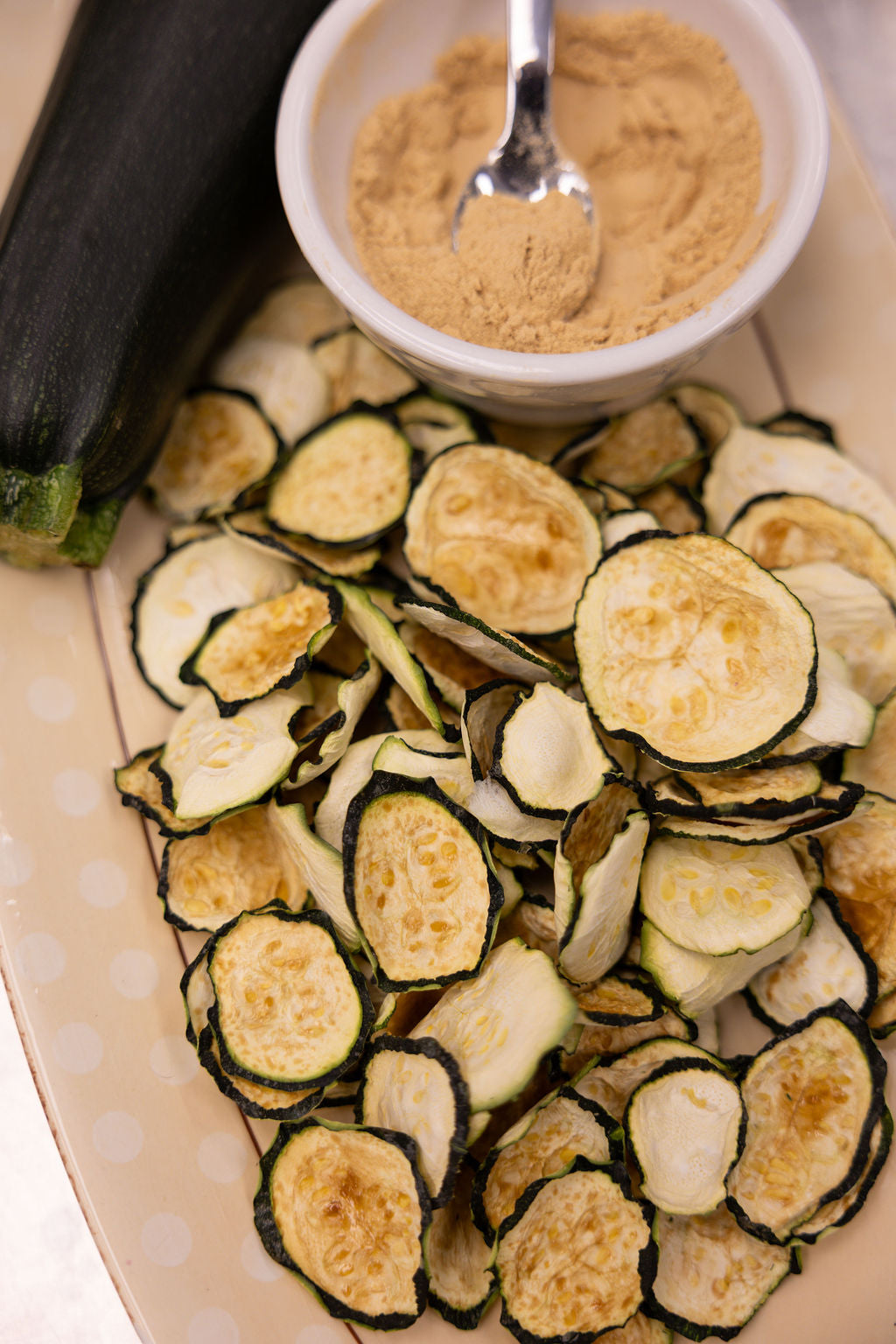 Zucchini chips on a plate with a bowl of seasoning and a zucchini next to them.