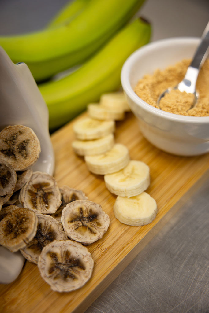 Banana slices, dried bananas, and a bowl of honey on a wooden board.